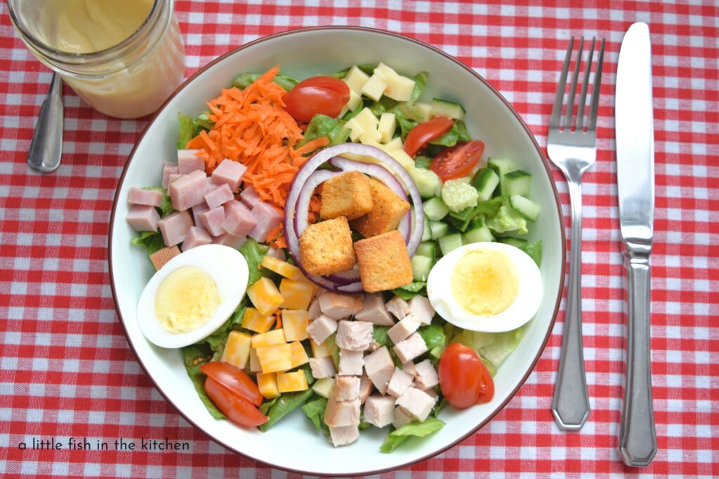 An eye-catching chef salad is served in a large, ceramic white salad bowl with a brown rim. The bowl is set atop a red and white checkered tablecloth. A fork and knife are set beside the bowl. Green, torn lettuce is topped with a rainbow of chopped raw veggies and diced deli meats and a sliced hard cooked egg.