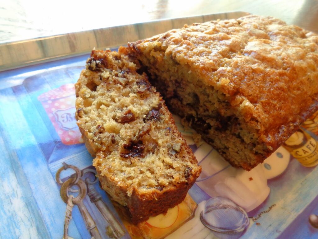 A fresh made loaf of banana bread is on a decorative serving board and ready to enjoy. The loaf is sliced showing a golden, tender crumb studded with melted chocolate chips and pecan pieces inside.