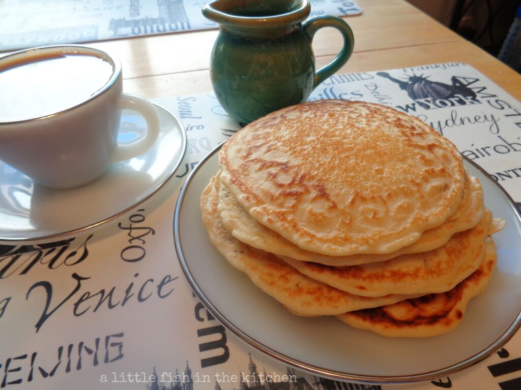 A stack of five fluffy homemade pancakes sits on a white plate, ready to serve. A cup of coffee sits next to the plate in a vintage Fire King cup and saucer set. A small teal ceramic pitcher sits behind the plate, it's filled with warm maple syrup for serving.