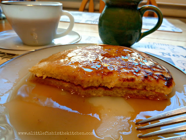 A white plate has two half eaten pancakes resting in a pool of maple syrup. The inside of the buttermilk pancakes looks tender, fluffy and tempting.