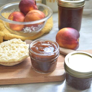 A small, clear glass mason jar filled with spiced peach butter sits on a wooded cutting board. The peach butter has a very smooth appearance and has a rich, dark golden color. Another small mason jar with a golden lid and rim is also filled with fruit butter is beside the cutting board. A sliced English muffin is on the other side of the cutting board. A golden, yellow kitchen towel sits behind the cutting board, a clear glass bowl filled with fresh peaches sits on it.