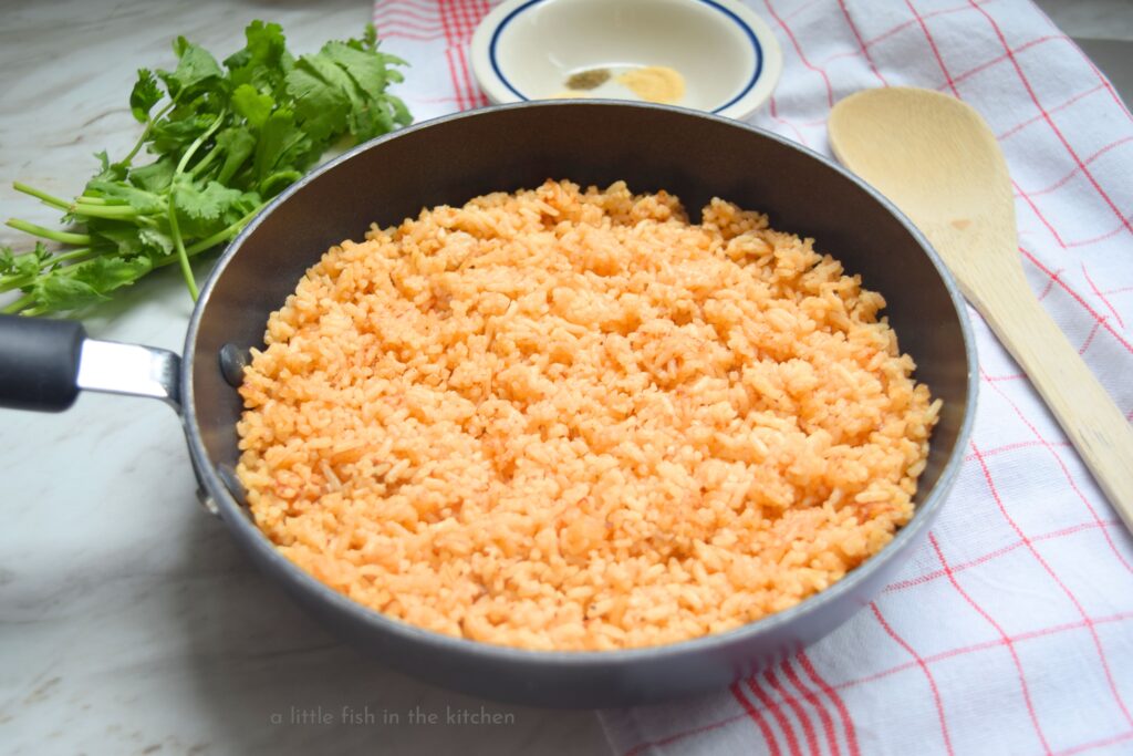A black metal skillet filled with freshly cooked Mexican rice rests on a red and white checkered kitchen towel. A wooden spoon is near the skillet for serving.