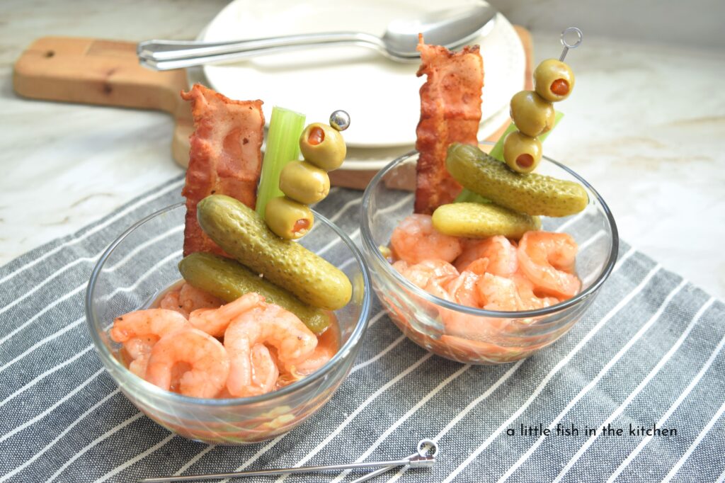 Two individual servings of eye-catching bloody Mary shrimp cocktail are ready to enjoy. The cooked shrimp are coated in a spicy red sauce on top of a bed of shredded lettuce. Each clear glass dish is garnished with single cocktail skewer loaded with green olives and mini gherkins. A single slice of crispy bacon and celery stick also adorn these tempting appetizers.