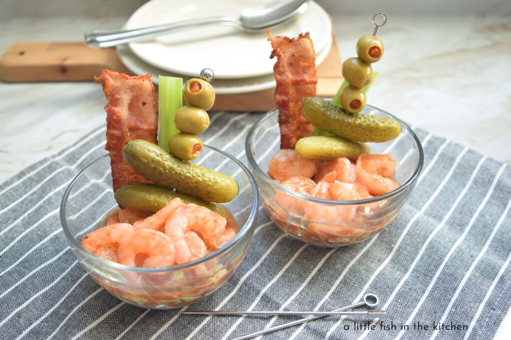 Two individual servings of eye-catching bloody Mary shrimp cocktail are ready to enjoy. The cooked shrimp are coated in a spicy red sauce on top of a bed of shredded lettuce. Each clear glass dish is garnished with single cocktail skewer loaded with green olives and mini gherkins. A single slice of crispy bacon and celery stick also adorn these tempting appetizers.