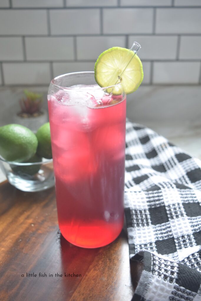 A sparkling red cranberry fizzer is in a clear glass high ball glass. The drink is a vibrant red color and the glass is garnished with a bright green fresh lime slice. A black and white checkered tea towel and a small bowl filled with fresh, whole limes are beside the drink.