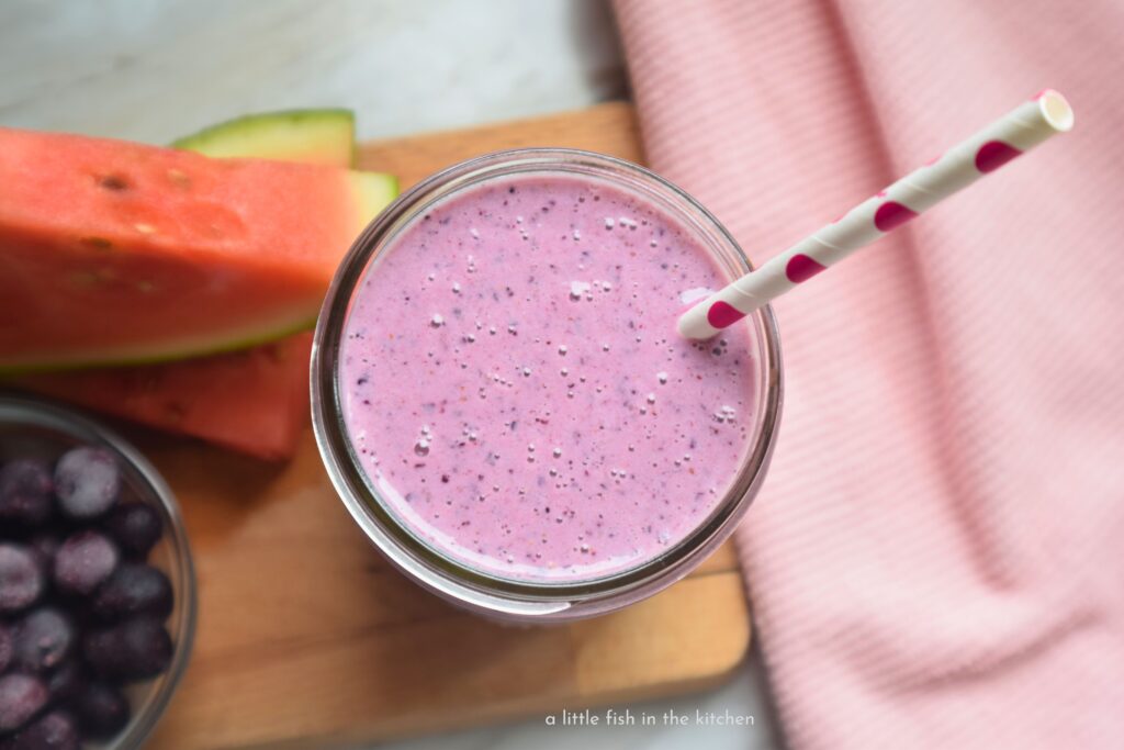A creamy purple smoothie is in a clear glass mason jar. Bits of blueberry skin and ground blue chia seeds are visible in this drink.  A light pink kitchen towel rests beside the smoothie.