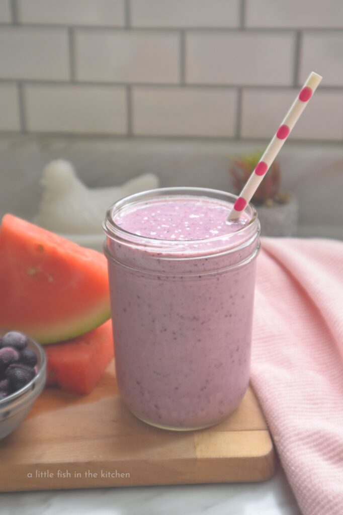 A creamy smoothie with a purple hue is in a clear mason jar accented with a white paper straw with pink polka dots. The glass is on a small wooden board, with a ramekin filled with frozen blueberries and watermelon. 
