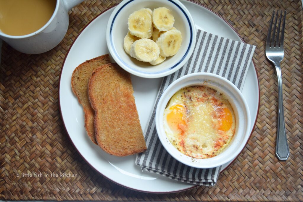 A white ceramic plate is filled with a small blue ramekin with two medium cooked eggs, a small bowl with fresh sliced banana, and a crisp slice of buttered wheat toast. Beside the place is a fresh cup of coffee with cream, and a fork is laying on the opposite side. Breakfast featuring tempting shirred eggs is served.