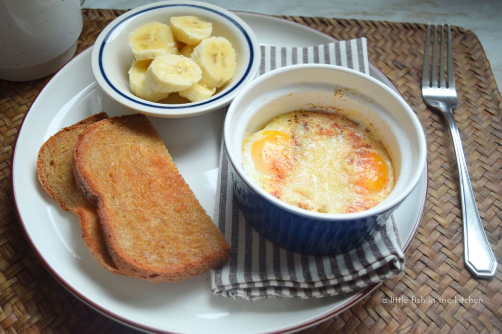 A white ceramic plate is filled with a small blue ramekin with two medium cooked eggs, a small bowl with fresh sliced banana, and a crisp slice of buttered wheat toast. Beside the place is a fresh cup of coffee with cream, and a fork is laying on the opposite side. Breakfast featuring tempting shirred eggs is served.