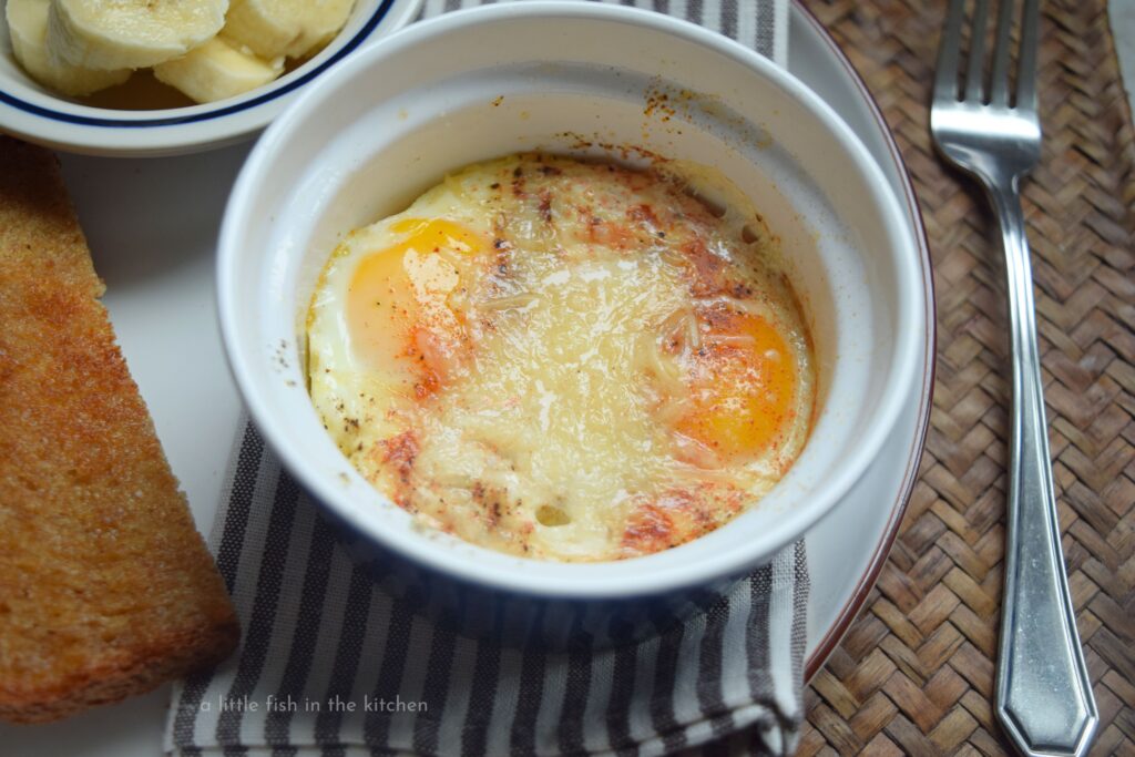 Two baked eggs are in a blue ceramic ramekin. The bright golden yolks look tempting, The melted parmesan cheese and colorful sprinkle of red smoked paprika make an eye catching presentation.