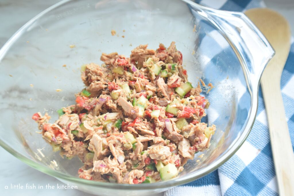 Freshly prepared Greek-style tuna salad is in a clear glass mixing bowl. Flaky tuna fish is speckled with chopped green cucumber, bits roasted red peppers and purple onions. It's colorful and inviting.