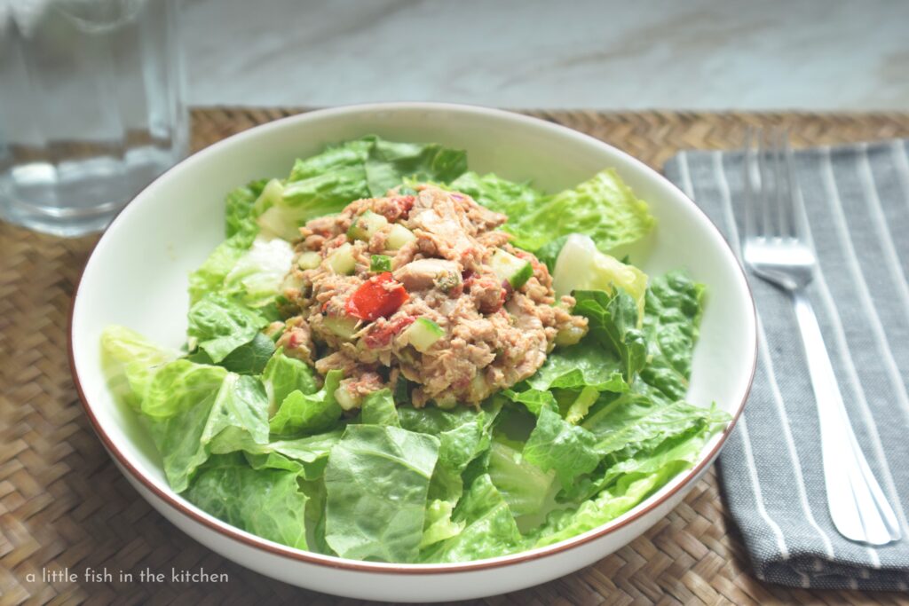 Colorful tuna salad with capers, red peppers and cucumbers sits on a bed of crisp, chopped Romaine lettuce and it's ready to enjoy. A gray cloth napkin with with white pin stripes and a fork on top, sits next to the bowl. 