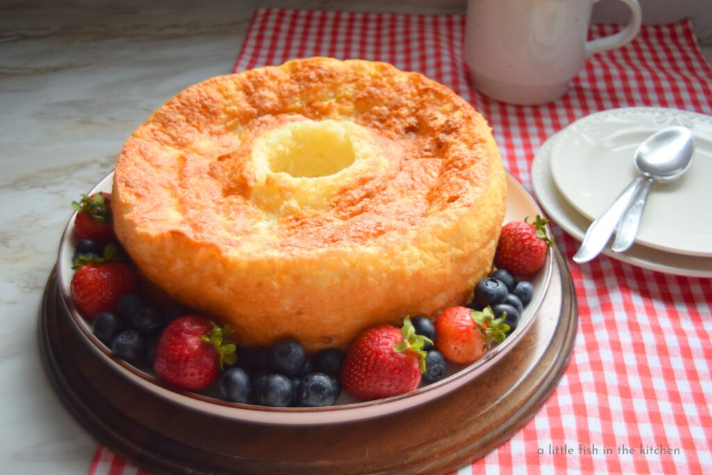A ring-shaped two-ingredient pineapple angel food cake is ready to serve and looking lovely surrounded by fresh summer berries on a large white plate. The plate sits on a small, round wooden cutting boars and the table is decorated with a red and white checker board runner. There are two, small white dessert plates, two spoons and a fresh cup of coffee nearby in the rustic scene.