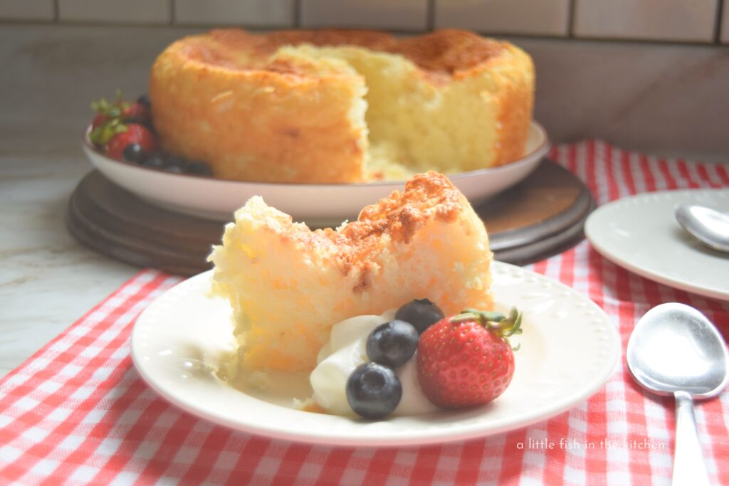 A slice of pineapple angel food cake is on a small white serving plate. It's garnished with a large dollop of whipped cream and a few fresh berries. The whole cake is slightly blurred in the background. 