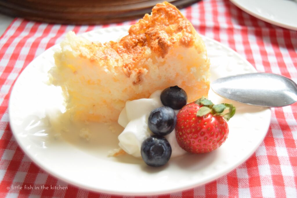 A single slice of this weight watcher's two ingredient angel food cake is on a white dessert plate and garnished with a fluffy dollop of whipped cream and summer berries. The cake looks irresistibly light and airy. with it's lovely golden hue, it's perfect for a summer dessert choice. 