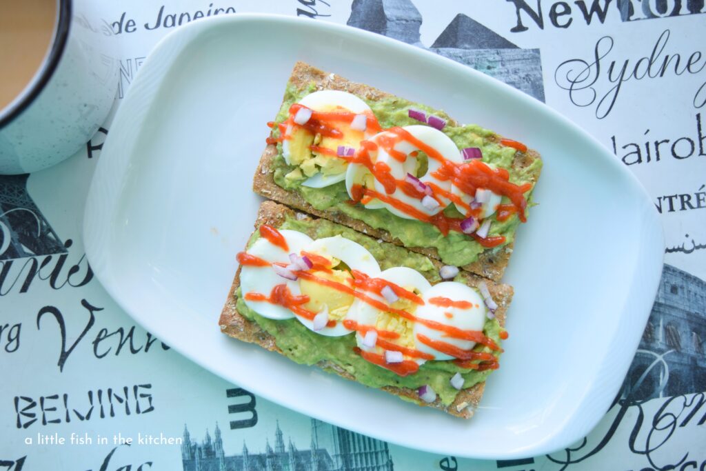 Two tempting pieces of Spicy Avocado and Egg Toast are ready to enjoy. The prepared toast is served on a small white platter. The green mashed avocado, white and yellow hard cooked egg, purple onions bits and a drizzle of red hot sauce is an eye-catching presentation.