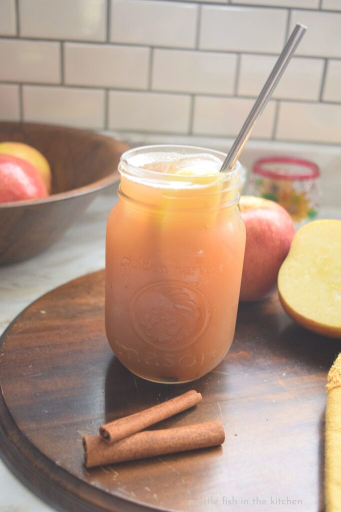 A clear glass mason jar is filled with golden Apple cider Arnold Palmer. The exterior of the mason jar looks chilled and cold and and apple slice is visible floating in the drink though the glass. A wooden bowl filled with ripe red and gold apples is slightly blurred in the background. 