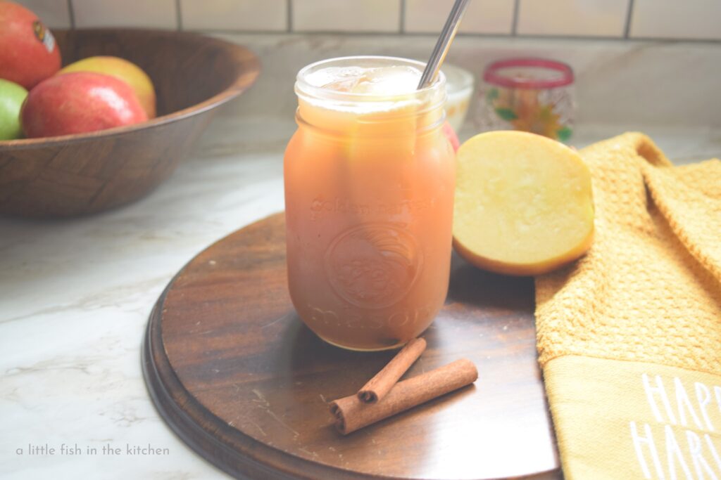A clear glass mason jar is filled with golden Apple cider Arnold Palmer. The exterior of the mason jar looks chilled and cold and and apple slice is visible floating in the drink though the glass. A wooden bowl filled with ripe red and gold apples is slightly blurred in the background. 