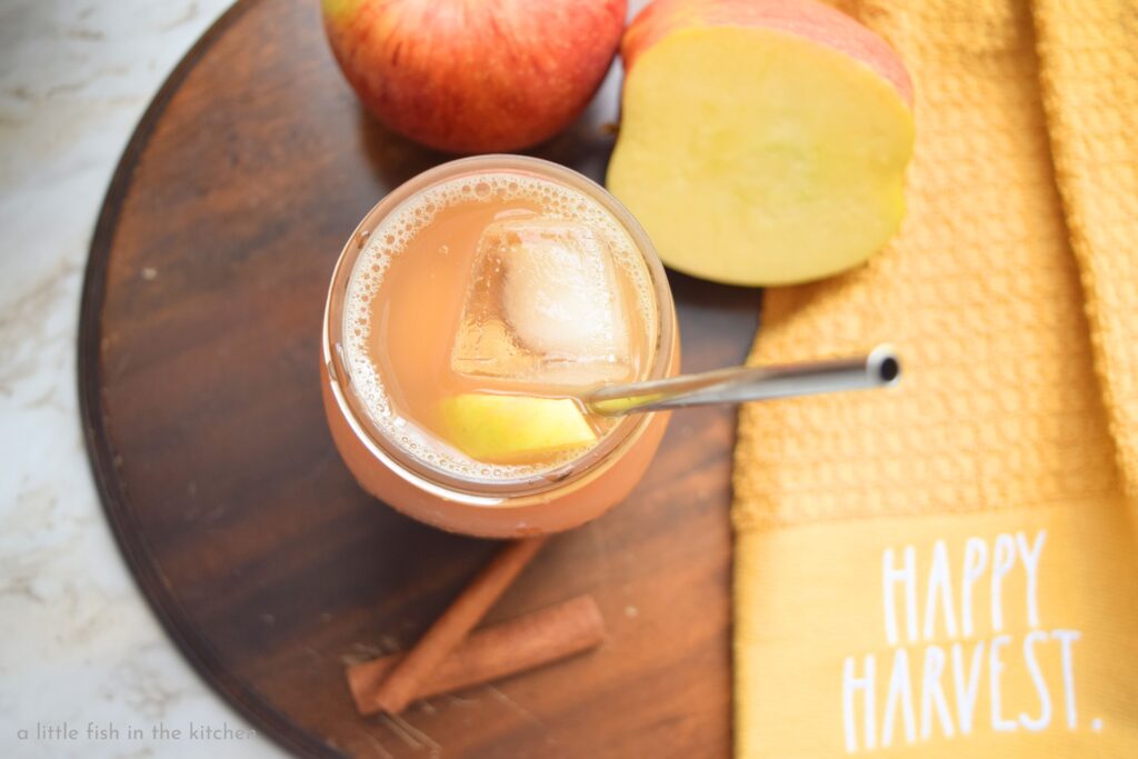 A golden colored combination of chilled apple cider and sweetened iced tea is in a mason jar served with a metal straw. The prepared drink rests on a small circular wooden cutting board. A gold-colored tea towel rests beside the mocktail, it's embroidered with the words "Happy Harvest"