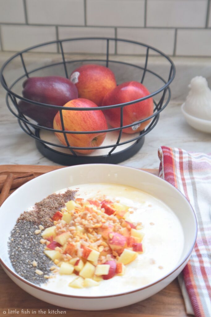 A bowl of spiced sweetened Greek yogurt is artfully topped with chia seeds, chopped apples, peanuts and toffee candy bits. The bowl is on a dark wooden cutting board with a red, orange and gold plaid tea towel laying nearby. A fruit bowl filled with red, ripe apples is slightly blurred in the background.