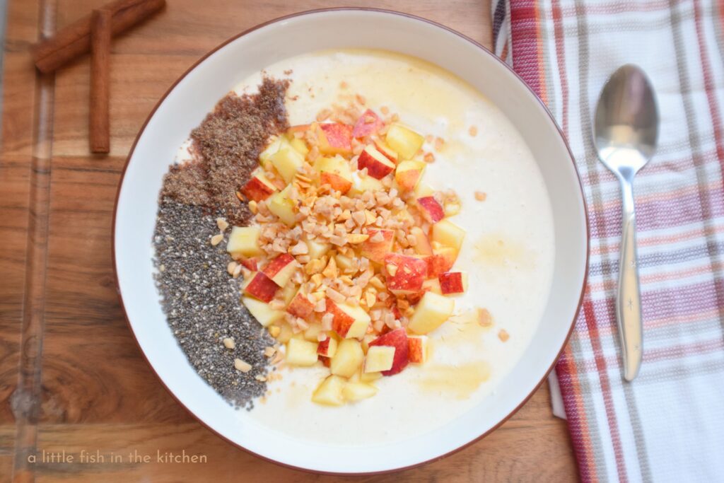 A bowl of spiced sweetened Greek yogurt is artfully topped with chia seeds, chopped apples, peanuts and toffee candy bits. The bowl is on a dark wooden cutting board with a red, orange and gold plaid tea towel laying nearby.
