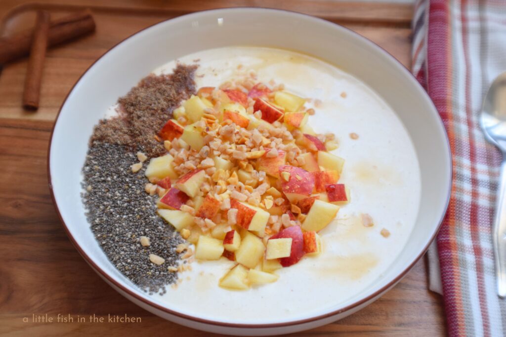 A bowl of spiced sweetened Greek yogurt is artfully topped with chia seeds, chopped apples, peanuts and toffee candy bits. The bowl is on a dark wooden cutting board with a red, orange and gold plaid tea towel laying nearby.