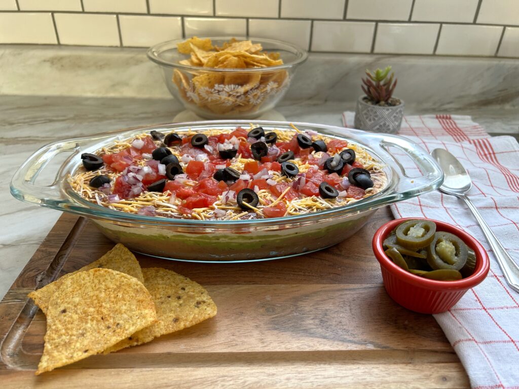 A colorful Mexican seven layer dip in a clear glass dish with layers of refried beans, seasoned sour cream, guacamole, salsa, shredded cheddar cheese, crisp lettuce, diced tomatoes, and sliced black olives, served with tortilla chips.