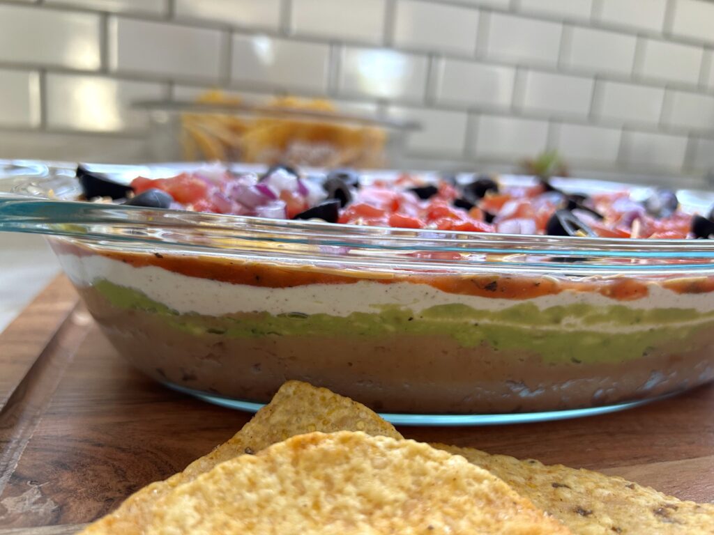 A colorful Mexican seven layer dip in a clear glass dish with layers of refried beans, seasoned sour cream, guacamole, salsa, shredded cheddar cheese, crisp lettuce, diced tomatoes, and sliced black olives, served with tortilla chips.