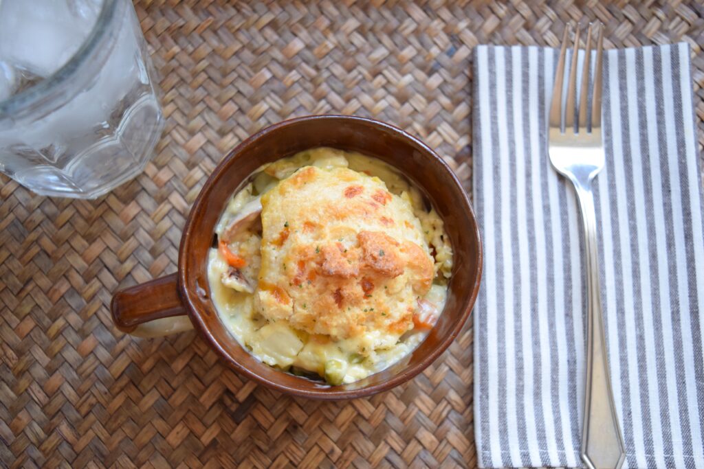 A single portion of this chicken cobbler is served in a rustic brown ceramic bowl. A chilled glass of water and a brown and white striped cloth napkin topped with a fork are next to the bowl. The bowl creamy cobbler is topped with a warm inviting buttery biscuit and bits of veggies and potatoes are visible in the rich soup filling. 