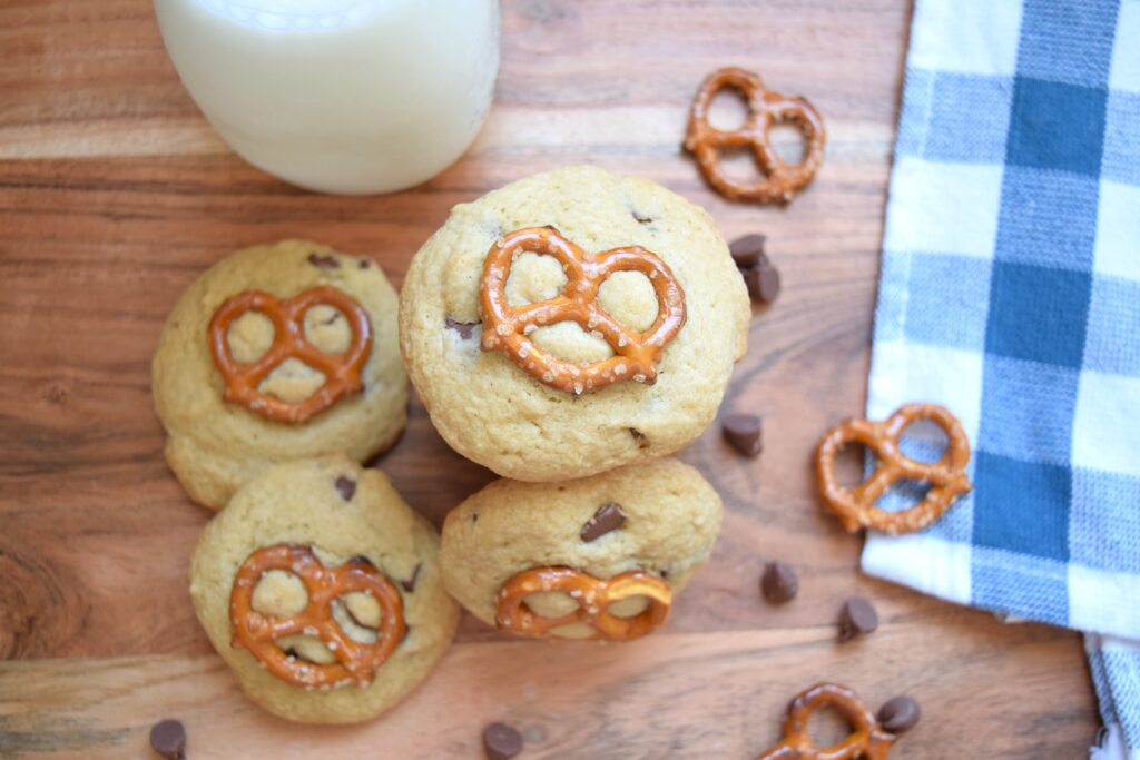 Tender, golden chocolate chip pretzel cookies are stacked and ready to served on a wooden cutting board. A blue and white tea towel is folded next to the warm cookies. Bits of melty chocolate chips are visible throughout the perfectly baked chocolate chip cookies and the toasted pretzel on top makes the sweet and salty treats extra tempting.