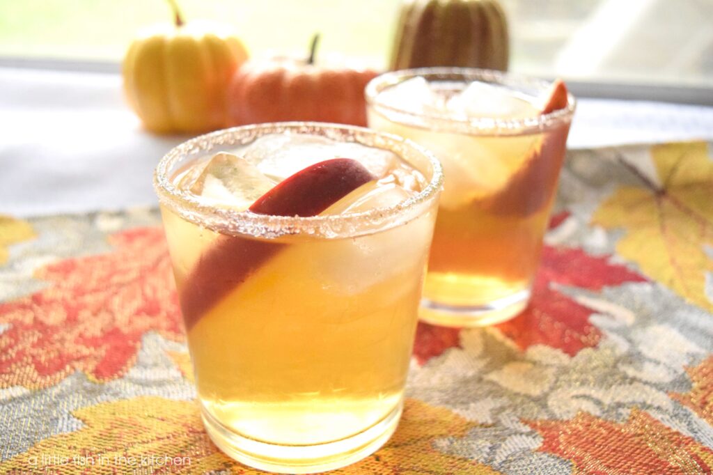 A bright golden beverage is served with a cinnamon and sugar rim as well as an apple wedge for garnish. The drinks are festively served on a sparkling silver table runner with orange, red, and gold leaves. 