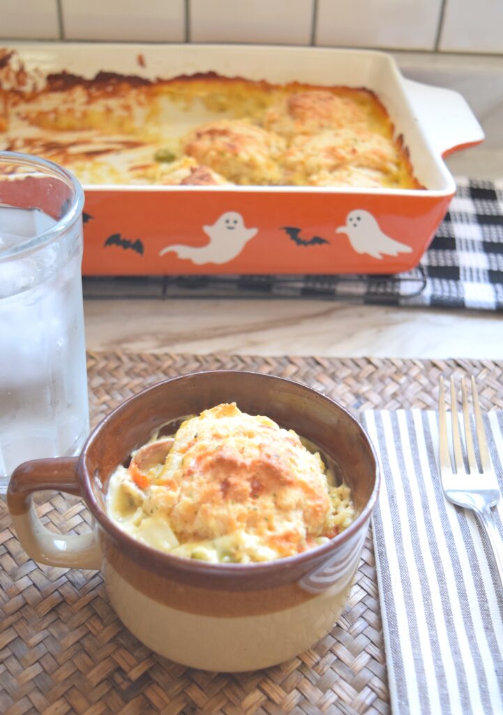 A single portion of this chicken cobbler is served in a rustic brown ceramic bowl. A chilled glass of water and a brown and white striped cloth napkin topped with a fork are next to the bowl. The bowl creamy cobbler is topped with a warm inviting buttery biscuit.