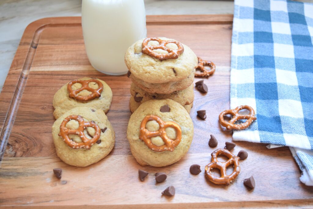 Perfectly golden chocolate chip cookies are baked to perfection with a mini pretzel baked into their tops. The cookies are eye-catching and with bits of gooey chocolate chips visible in the tender cookies. The cookies are served with milk on a wooden cutting board. A blue and white checkered kitchen towel is neatly folded off to the side.