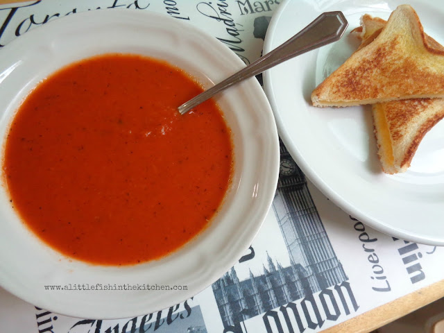 Creamy tomato soup is served in a white ceramic bowl, there is a spoon resting in the bowl with the soup. It has a rich, deep red color and it looks flavorful. Tiny bits of cracked black pepper and herbs are visible in the soup. A white plate with a buttery grilled cheese sandwich sits beside the bowl of soup.  
