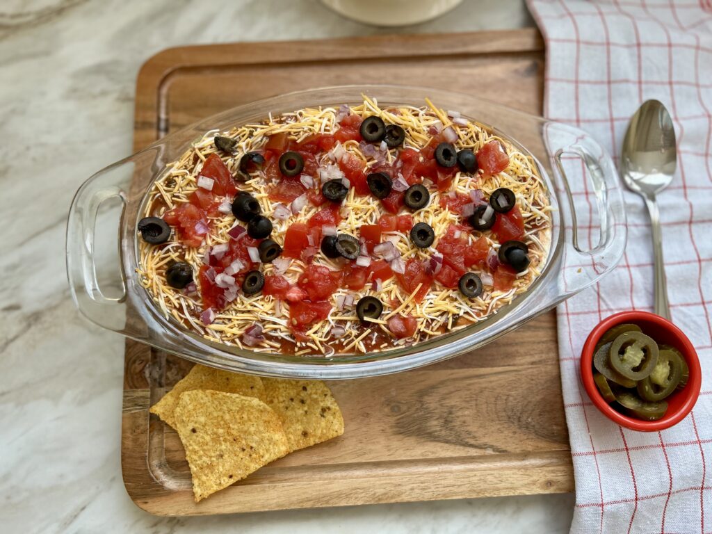 The colorful layers of a classic 7-layer dip are finished with a generous blanket of shredded cheddar cheese, juicy diced tomatoes, sliced black olives, and finely diced red onion creating a beautiful contrast of colors and textures.