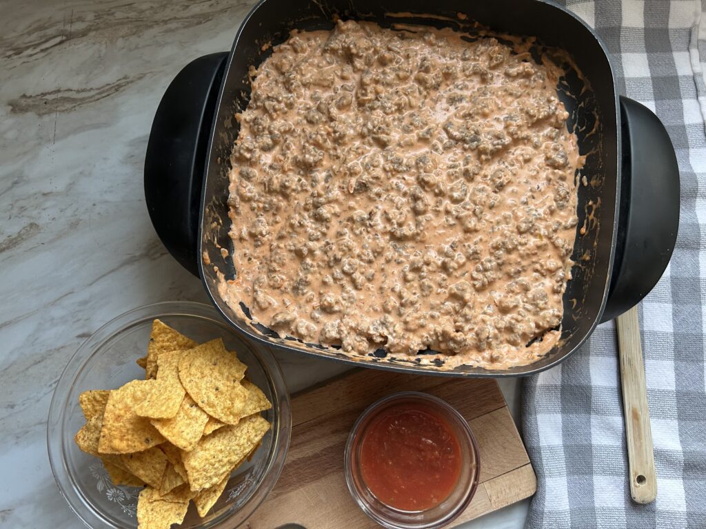 A black electric skillet is warming a creamy dip, a glass bowl with tortilla chips and a small ramekin with red salsa are next to the skillet. The warm cream cheese dip has a pinkish color and bits of browed sausage are visible thoughout the mix. A gray and white checked tea towl and wooden spoon are laying next to the skillet on the right side. 