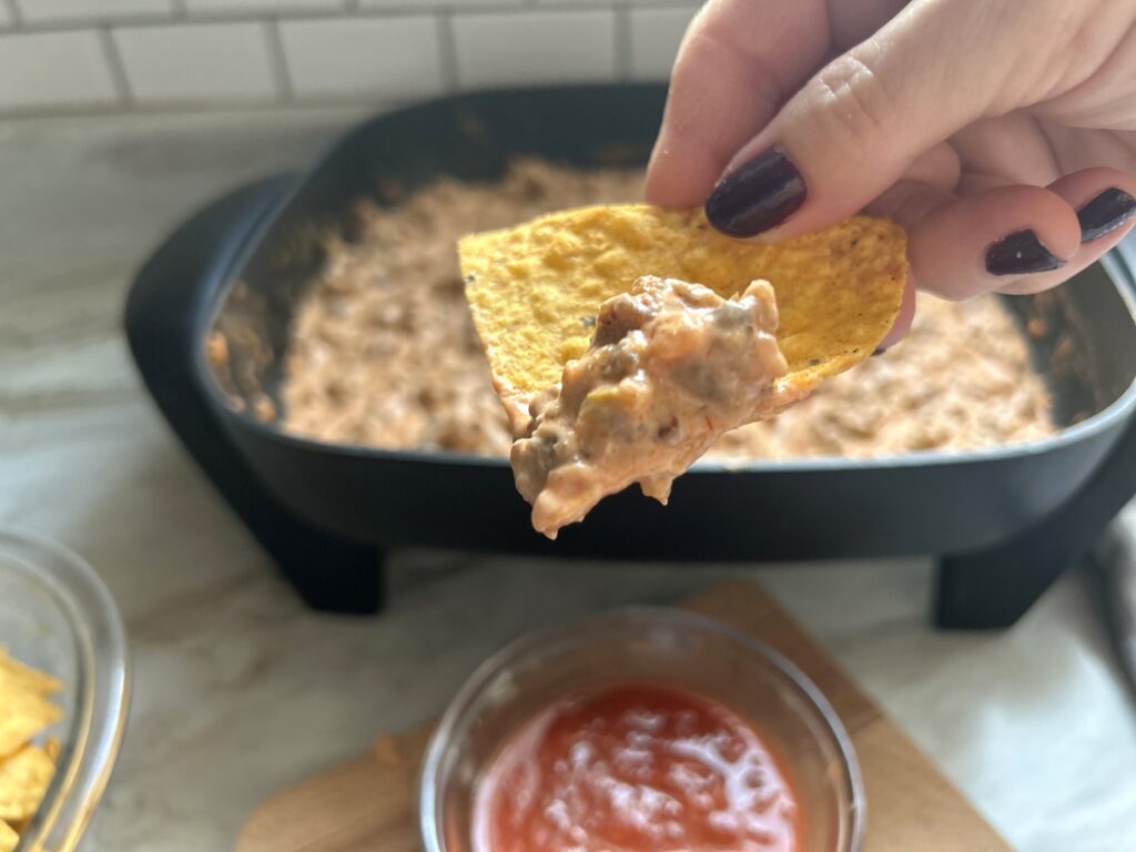 A hand holds a chip covered in warm cream cheese queso. The dip looks creamy, hearty and inviting with bits of sausage and spices visible in the dip. 