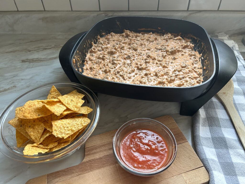 A black electric skillet is warming a creamy dip, a glass bowl with tortilla chips and a small ramekin with red salsa are next to the skillet. The warm cream cheese dip has a pinkish color and bits of browed sausage are visible thoughout the mix. A gray and white checked tea towl and wooden spoon are laying next to the skillet on the right side. 
