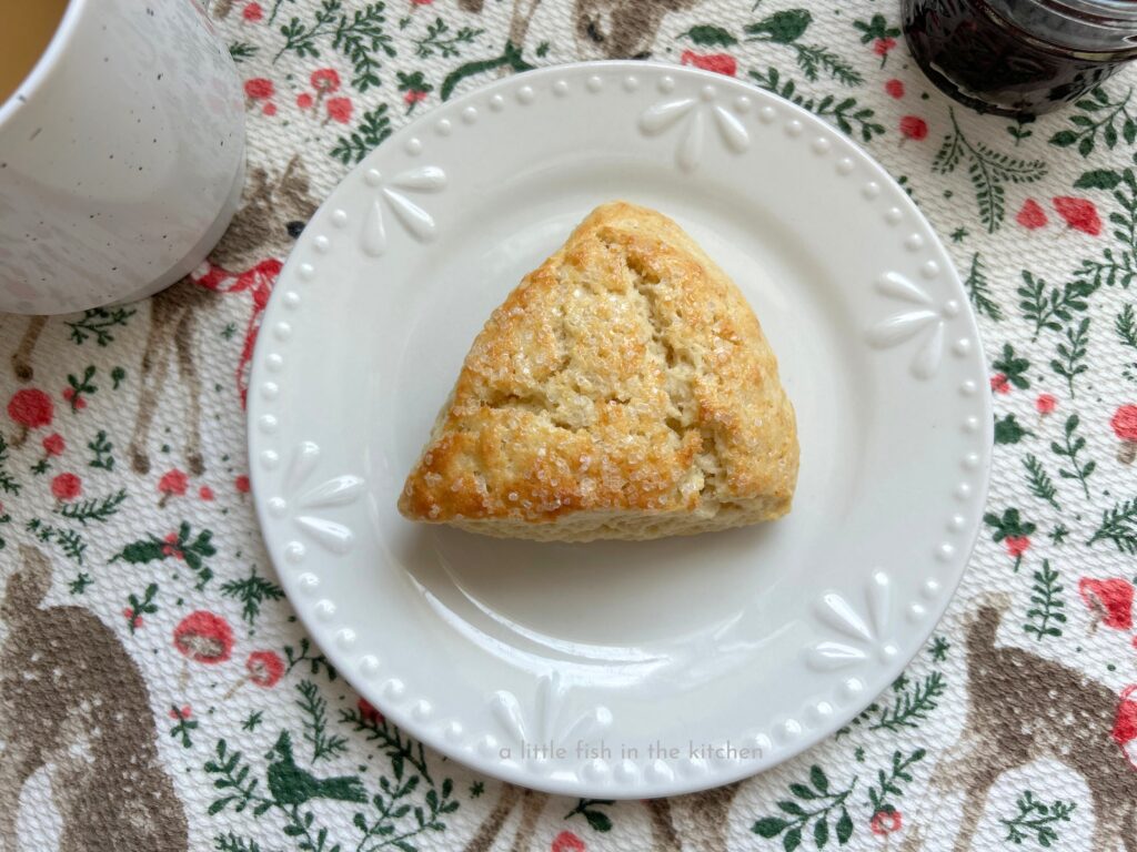 A single flakey whipping cream scone is served on a small white ceramic dessert plate. The top is warmly toasted and crunchy bits of white sparkling sugar are visible on the lightly crackled top.A white tea towl with a bright pattern of greenery, green birds, deer and red and white small mushrooms is underneath the metal baking pan. The colorful towel gives the photo a festive appearance.