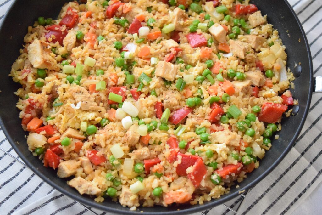 Colorful cauliflower fried rice is ready to serve in a black skillet. The skillet is on a rack and a white tea towel with black stripes. The colorful ingredients: red bell pepper, green onions, and peas and carrots give this healthful dinner a confetti like appearance.