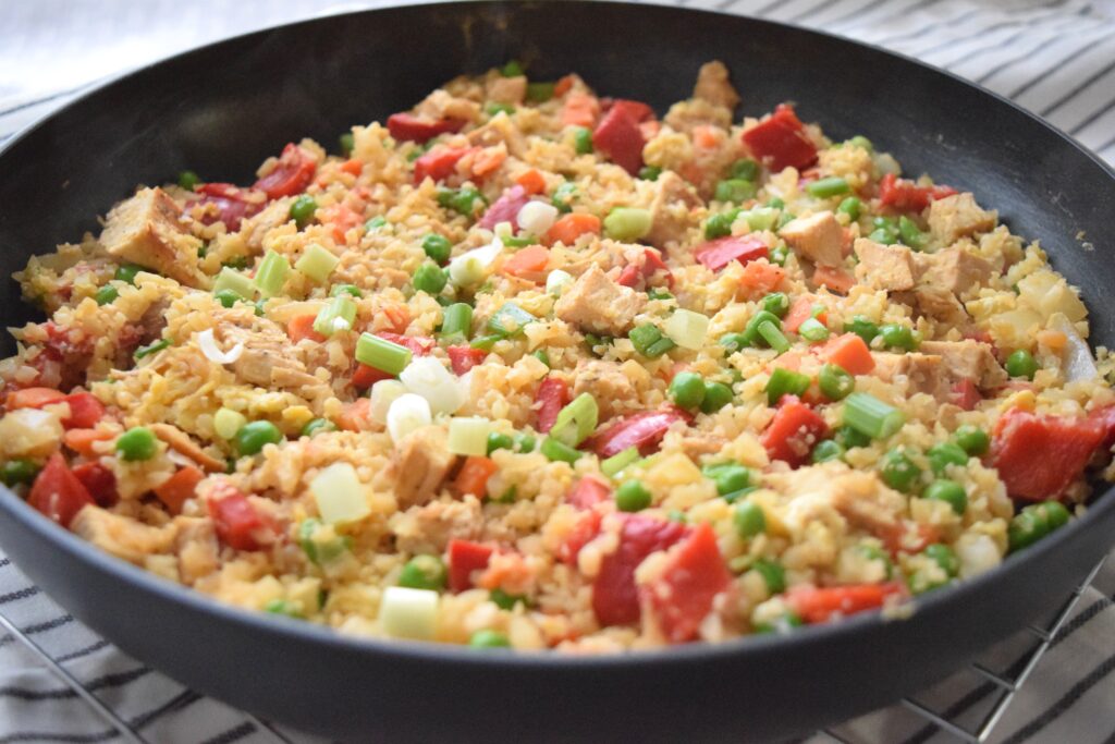 Colorful cauliflower fried rice is ready to serve in a black skillet. The colorful ingredients: red bell pepper, green onions, cooked bits of chicken and peas and carrots give this healthful dinner an inviting confetti like appearance.