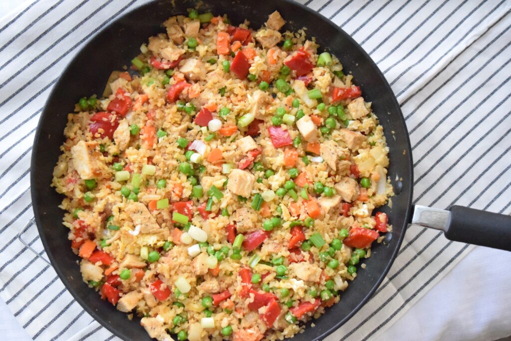 Colorful cauliflower fried rice is ready to serve in a black skillet. The colorful ingredients: red bell pepper, green onions, and peas and carrots give this healthful dinner a confetti like appearance.