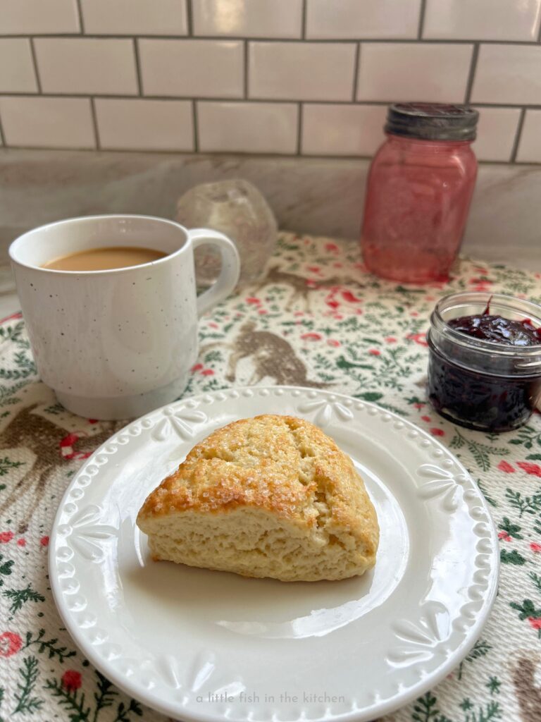 A single whipping cream scone is served on a small white ceramic plate. The top is lightly browned, toasted and bits of white sparkling sugar are visible on the lightly crackled top. A small glass mason jar with dark red jam is next to the plate and a white coffee cup filled with coffee is on the other side. A white tea towl with a bright pattern of greenery, green birds, deer and red and white small mushrooms is underneath the metal baking pan. The colorful towel gives the photo a festive appearance.