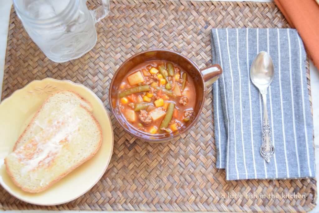 A brown ceramic bowl with a handle is filled with old-fashioned hamburger vegetable soup with v8 juice. The amber-colored broth is studded with bits of tender ground beef, corn, peas, carrots, cut green beans and diced potato. A gray cloth napkin with white stripes is topped with a spoon to the right side of the bowl. A small yellow plate with two pieces of homemade buttered bread is to the left of the bowl of soup. The meal is served on a vintage woven wood place mat. 