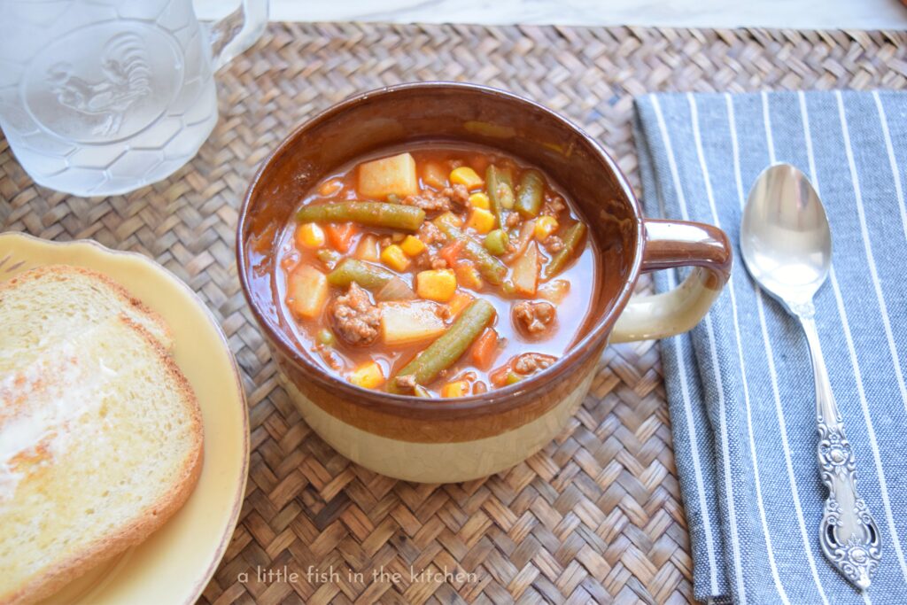 A vintage brown ceramic bowl with a handle is filled with old-fashioned hamburger vegetable soup made with v8. The amber-colored broth is studded with bits of tender ground beef, corn, peas, carrots, cut green beans and diced potato. A soup spoon and a gray napkin with white stripes is near the hearty bowl of ground beef vegetable soup. 