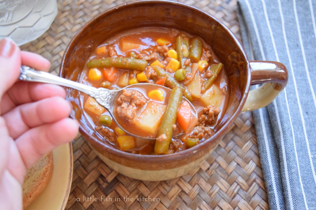 A hand holds a spoon filled with  hearty hamburger soup with bits of diced potato, tender ground beef, corn, green beans and carrots mixed in. 