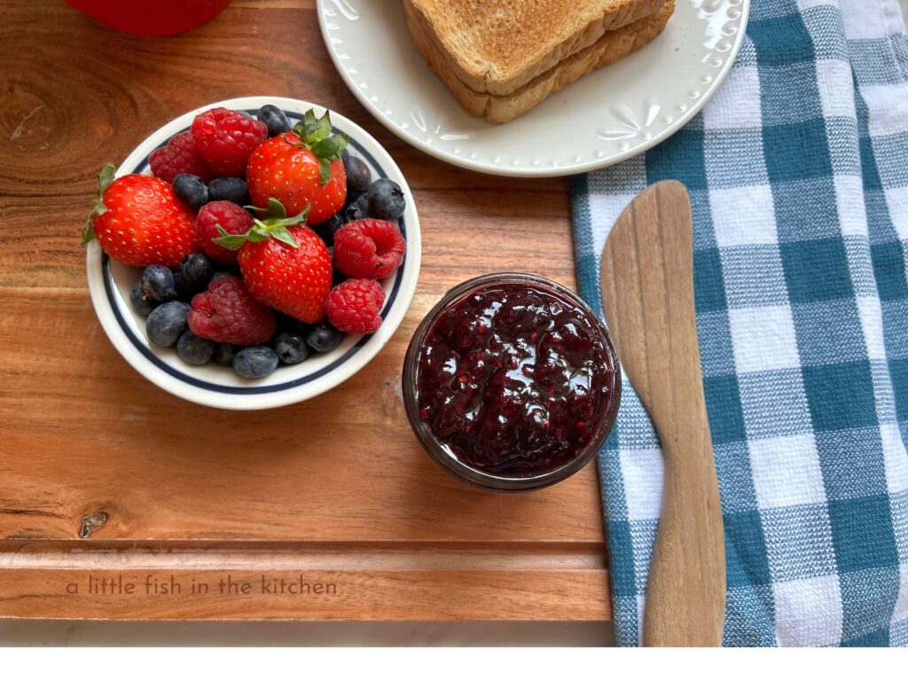 A small white ceramic ramekin with a blue line around the trim is filled with a mix of fresh blueberrie, raspberries and strawberries. Next to the ramekin is a small clear glass mason jar filled with a vibrantly violet-colored mixed berry jam. A wooden butter knife laying on a blue and white checkered is next to the jar. Directly behind the jar jam jar, sits a small white plate with two pieces of plain toast. All of these items are arranged on a large wooden cutting board giving the meal setting a very rustic, country appearance.