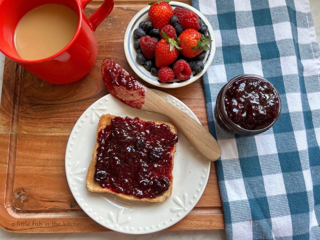 A small white ceramic ramekin with a blue line around the trim is filled with a mix of fresh blueberrie, raspberries and strawberries. Next to the ramekin is a small clear glass mason jar filled with a vibrantly violet-colored mixed berry jam. A small white plate serves a single slice of toast that's generously slathered with fresh mixed berry jam. A small wooden butter knife, the top end covered in jam, rests on the edge of the plate. 