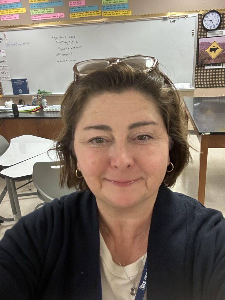 A selfie picture of myself, at work as a teacher. I'm in the center of a classroom with a large dry erase board in the background. I'm wearing a blue cardigan and I have my reading glasses on the top of my head. I am smiling and happy to be near the end of the work day!