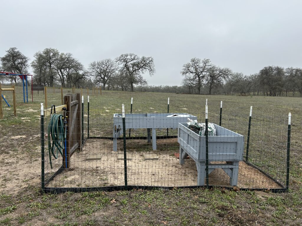 Small enclosed garden area with a garden hose rolled up on the side of the entry door. Two homemade raised beds are painted with light gray paint are enclosed by fencing. Bags of dirt at inside the beds. Soon they will be ready for planting.